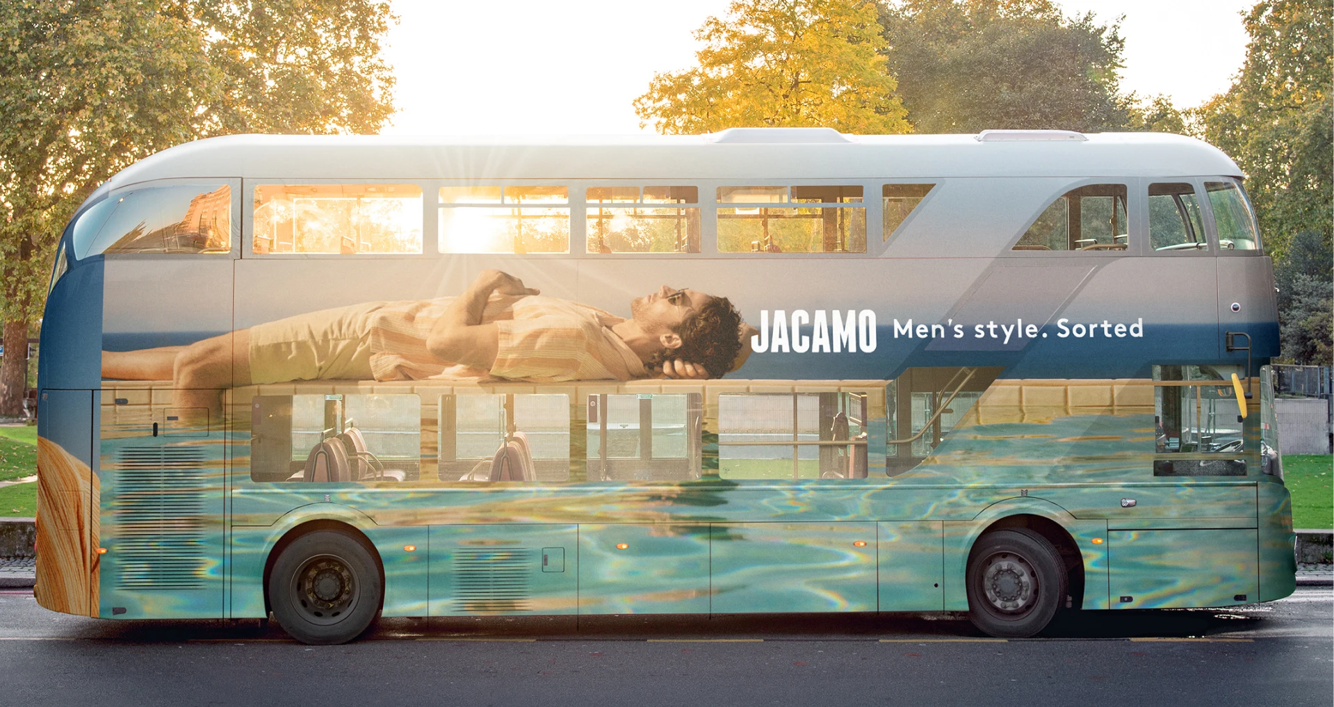 Double-decker bus wrapped with a Jacamo men’s fashion advertisement showing a man reclining above a water-themed graphic, parked along a tree-lined street.